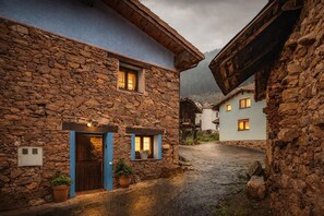 A stone building with a wooden door and windows, a paved courtyard, and another building in the background. - Apartment 'El Ablanu, Hot Tub & Mountain Views' with Terrace and Wi-Fi (Proaza)