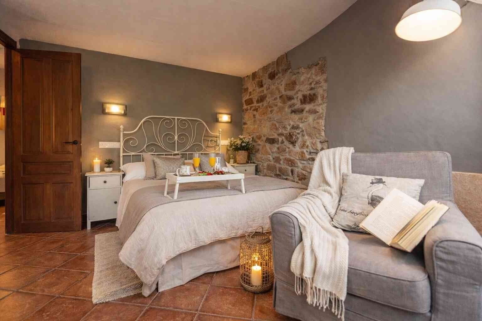 A bedroom with a stone wall, a bed with a headboard, bedside tables with lamps, a grey armchair, and a bedside table with a tray of breakfast items.
