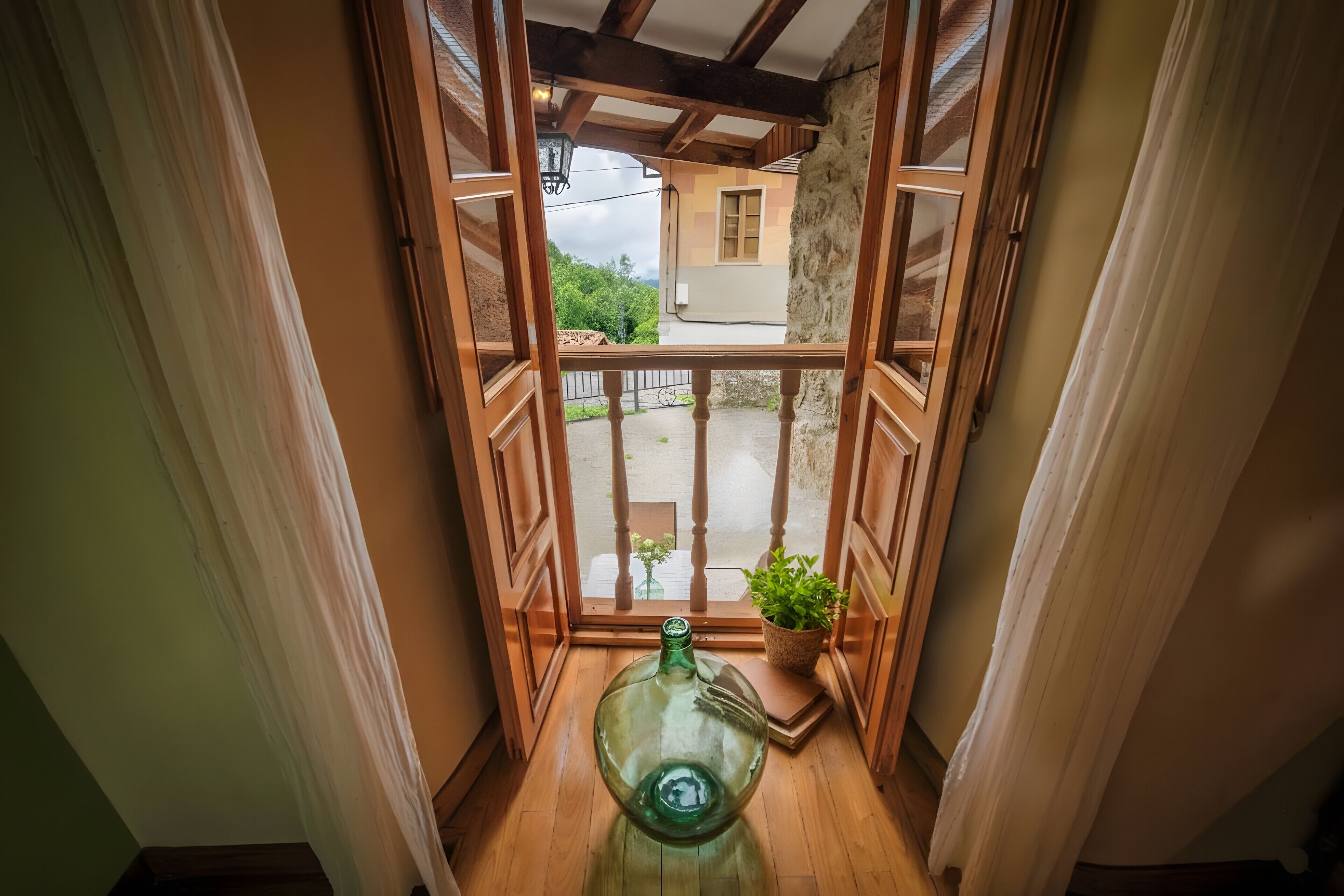Una habitación con suelo de madera, un ventanal con vistas a un edificio y vegetación, y una botella de vidrio verde sobre el suelo.