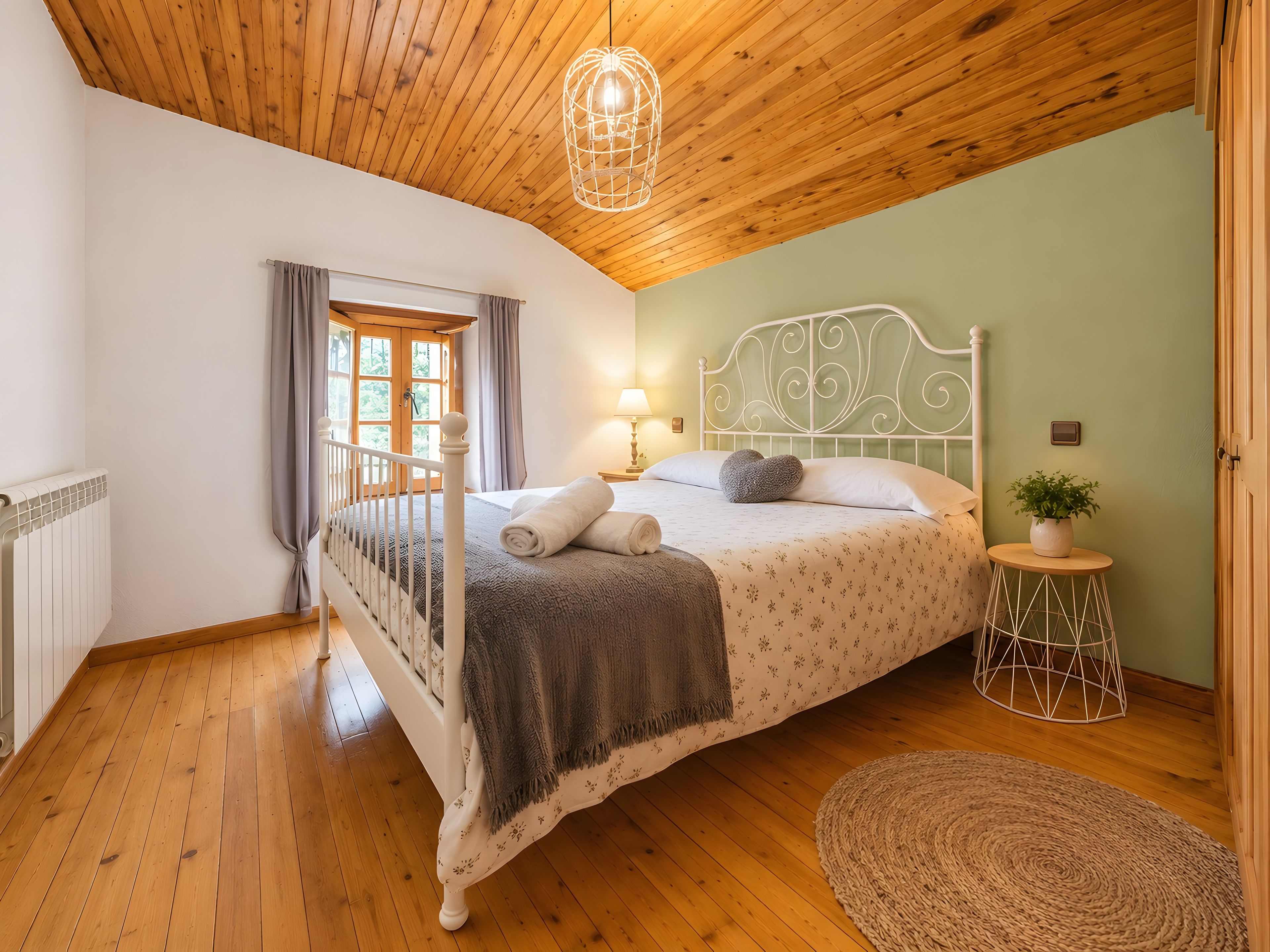 Quarto com teto de madeira, cama com cabeceira branca, mesa de cabeceira com abajur e janela com cortinas.