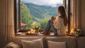 Una persona sentada junto a una ventana con vistas a un paisaje montañoso, sosteniendo una taza.