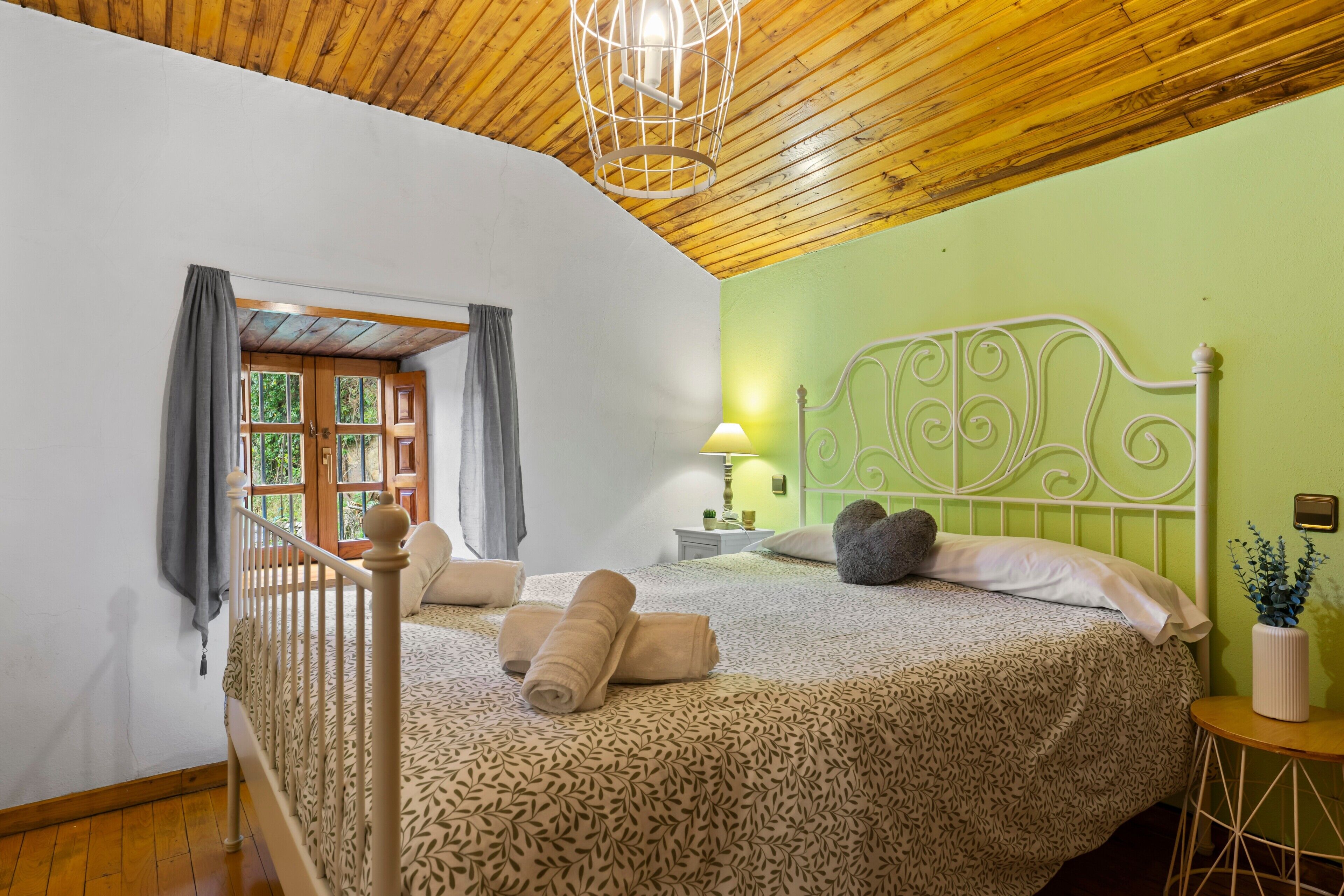 Un dormitorio con techo de madera, cama con cubrecama estampado, mesita de noche con lámpara y vistas a través de una ventana con cortinas.