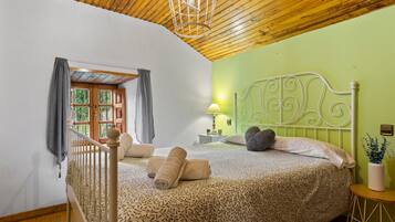 Un dormitorio con techo de madera, cama con cubrecama estampado, mesita de noche con lámpara y vistas a través de una ventana con cortinas.