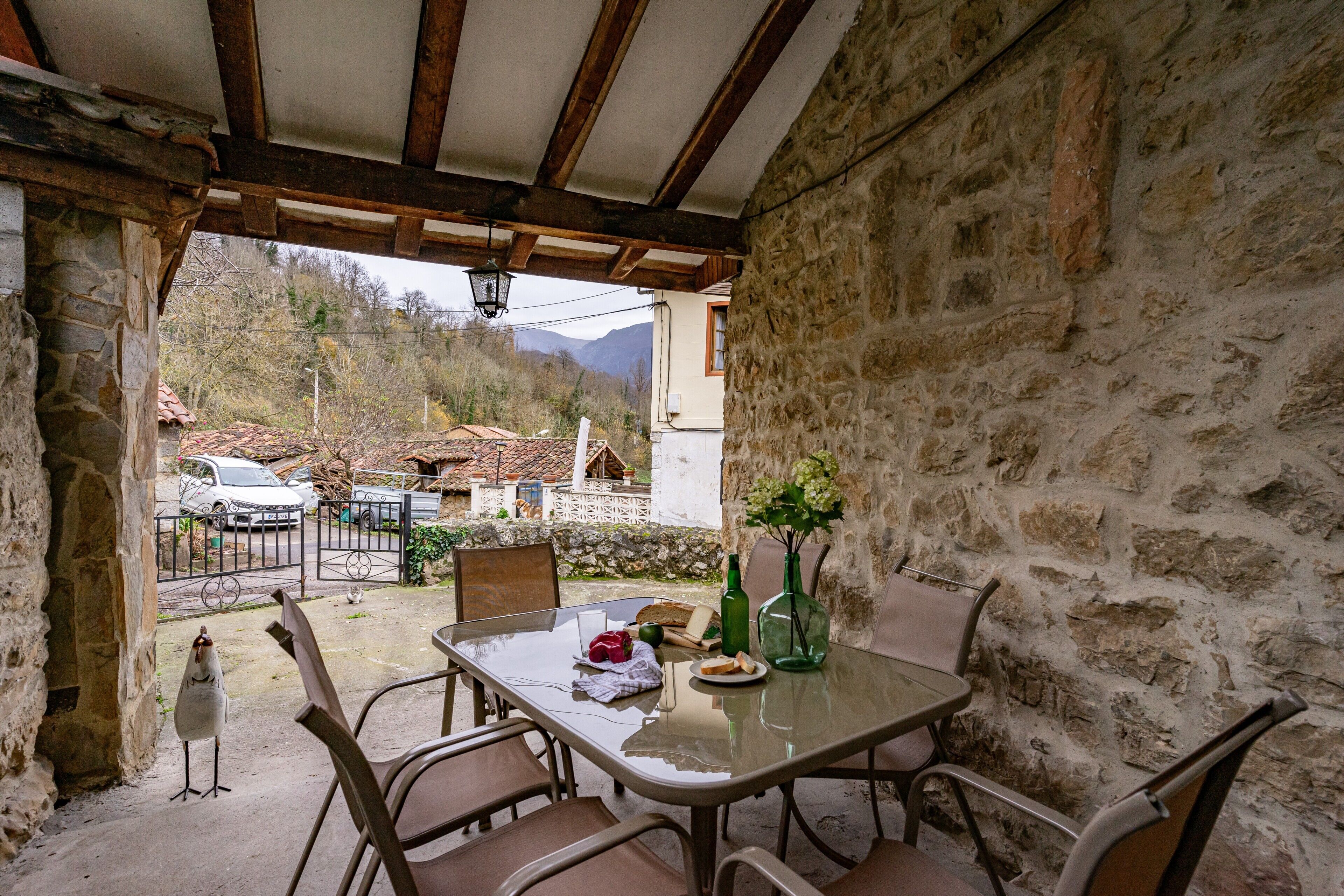 Un porche cubierto con paredes de piedra, una mesa con sillas, un pájaro y vistas a un pueblo.