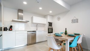 Une cuisine moderne avec des meubles blancs, une hotte en acier inoxydable et un coin repas comprenant une table en bois et des chaises blanches.