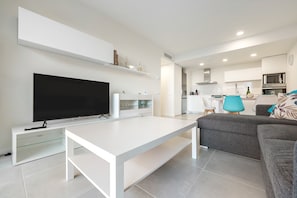 Un salon moderne avec un téléviseur à écran plat, un meuble de rangement blanc, un canapé gris et un coin cuisine à l’arrière-plan.