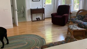 A spacious living room with hardwood flooring, a large window with curtains, a ceiling fan, and a door leading to another room.