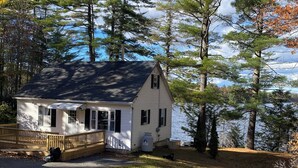 A small, single-story house with a porch, surrounded by trees and a body of water.