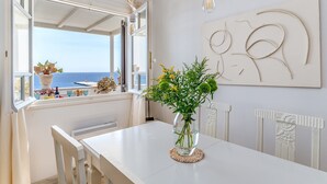 A dining area with a table, chairs, a vase with flowers, a view of the sea, and a wall with abstract art.