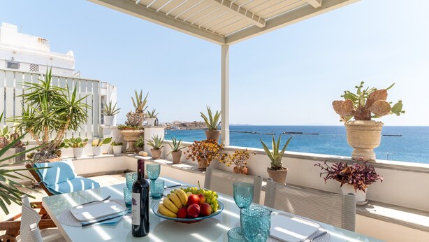 A table set with fruits, a bottle, and blue glassware on a balcony with potted plants and a view of the sea.