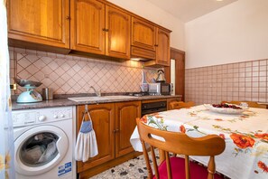 Une cuisine avec des meubles de rangement en bois, un lave-linge, un four à micro-ondes et une table à manger recouverte d’une nappe à motifs floraux.