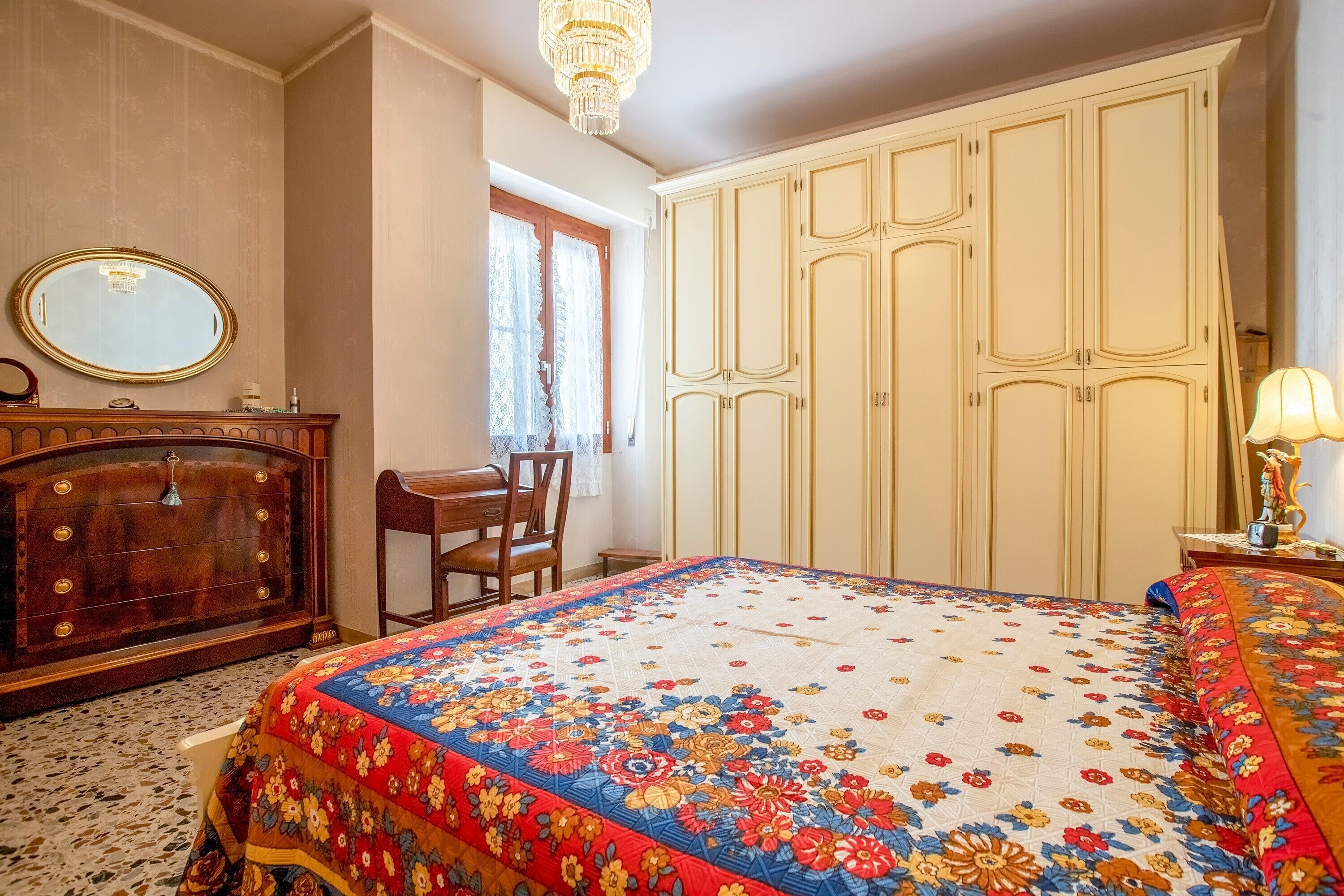 A bedroom with a floral bedspread, a wooden dresser, a chair, a wardrobe, and a chandelier.