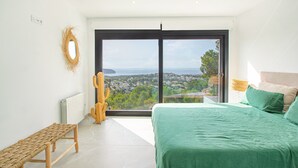 A bedroom with a large window, a bed, a bench, and a cactus.