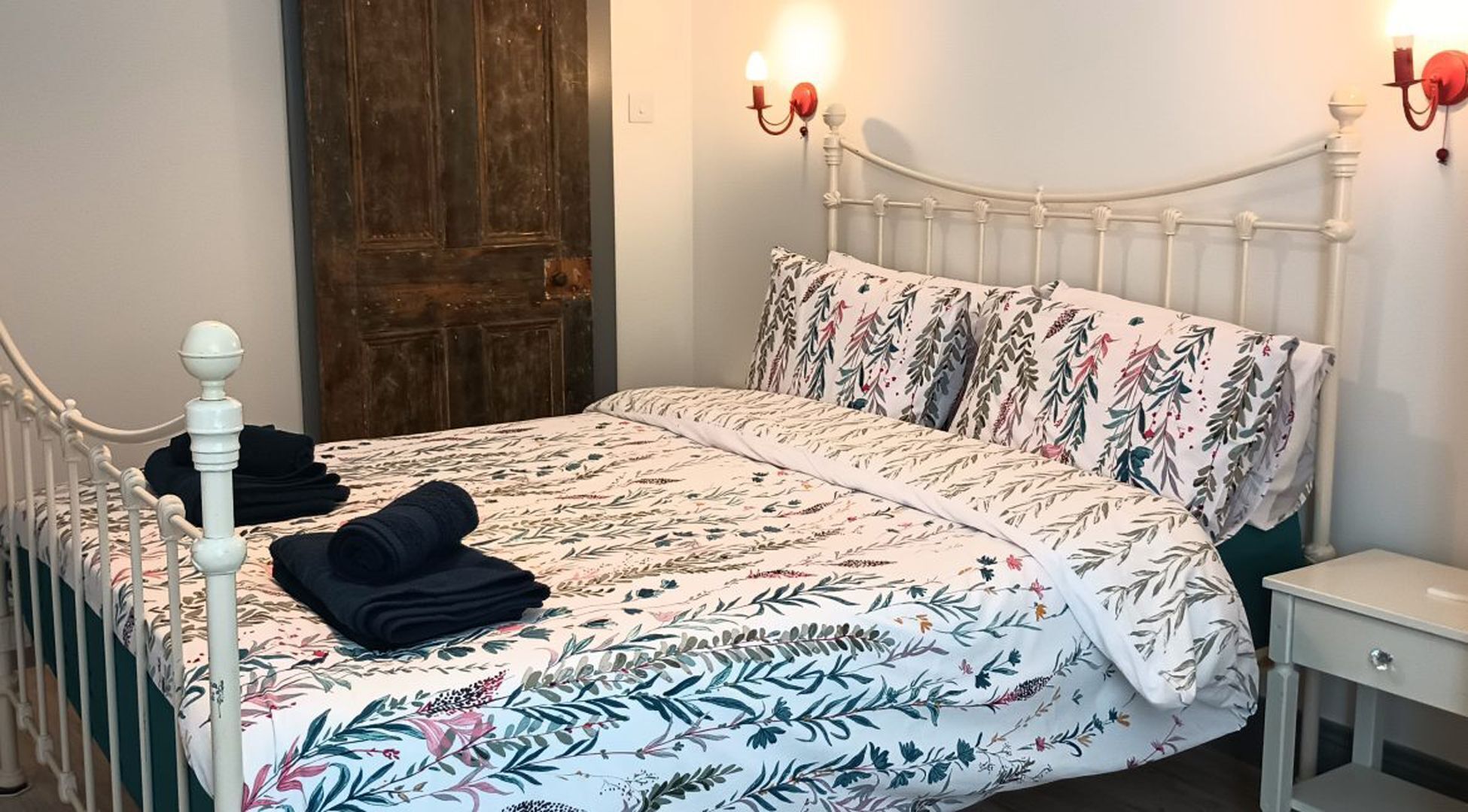 Een bed met een bloemenprint deken, een houten deur en twee wandlampen.