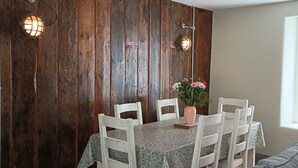A dining area with a rustic wooden wall, white chairs, a table with a floral tablecloth, and a vase with flowers.
