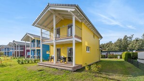 Una casa amarilla con balcón, rodeada de un césped y otras viviendas.