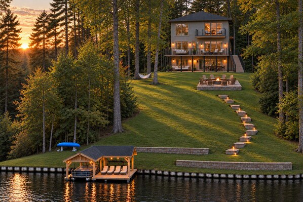A two-story house with a deck and outdoor seating area, surrounded by trees and a body of water.