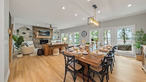 A spacious dining area with a wooden table set for six, surrounded by black chairs. A stone fireplace and a large window with a view of the outdoors are visible. - Luxe Lakefront: Spa, Kayaks, Pickleball, & Theater (Macon)