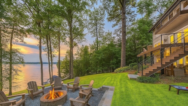 A lakeside outdoor seating area with Adirondack chairs and a fire pit, overlooking a body of water and a modern building with a staircase. - Luxe Lakefront: Spa, Kayaks, Pickleball, & Theater (Macon)