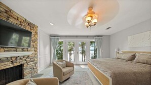 A bedroom with a stone fireplace, a bed, a TV, and a view of the outdoors. - Luxe Lakefront: Spa, Kayaks, Pickleball, & Theater (Macon)
