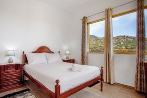 Une chambre d’hôtel avec un lit en bois, deux tables de chevet avec des lampes et une vue sur un paysage vallonné, visible à travers une grande fenêtre.