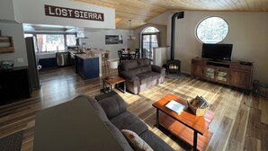 A spacious living area with a wooden ceiling, a fireplace, a television, and a kitchen area visible through an archway.