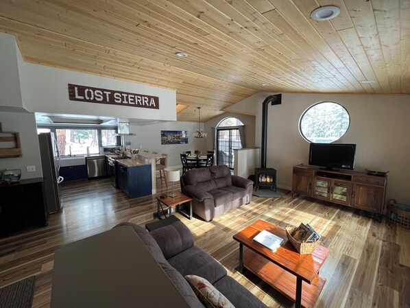 A spacious living area with a wooden ceiling, a fireplace, a television, and a kitchen area visible through an archway.