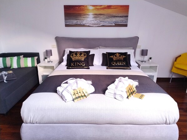 A neatly made bed with pillows labeled 'KING' and 'QUEEN,' a bedside table with a lamp, a chair, and a painting of a sunset over the ocean on the wall.