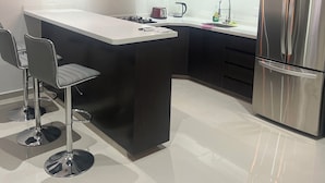 Une cuisine moderne avec des meubles de couleur sombre, un réfrigérateur en acier inoxydable, un îlot central doté de tabourets et un éclairage suspendu.