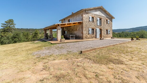 Una casa de piedra con un porche cubierto, camino de grava y vista a la colina.