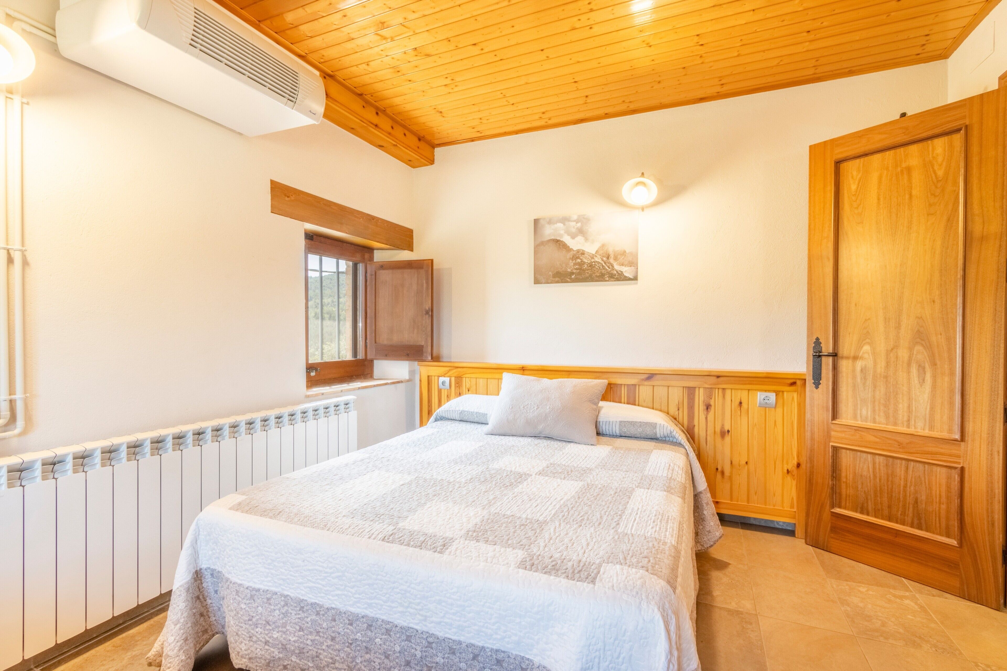 A bedroom with a wooden ceiling, a radiator, a bed with a quilt, a wooden door, and a wall-mounted light fixture.