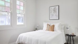 A minimalist bedroom with a large window, a bed with white bedding, two bedside tables with lamps, and a framed abstract painting on the wall. - Austin Cottage - Pet friendly holiday house in Newcastle (The Junction)