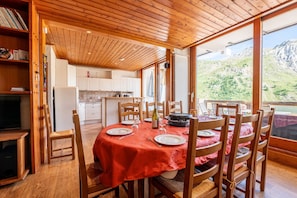 A dining area with a wooden table set for six, a kitchen in the background, and a view of a mountainous landscape through large windows. - Apartment 'Shamrock 2B' with Mountain View, Balcony and Wi-Fi (Tignes)