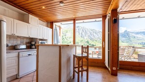 A kitchen with wooden cabinets, a dishwasher, and a view of mountains through large windows. - Apartment 'Shamrock 2B' with Mountain View, Balcony and Wi-Fi (Tignes)