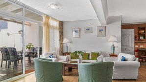 Una sala de estar con puerta de vidrio que da a un espacio exterior, un sofá blanco, sillones verdes y una mesa de centro.