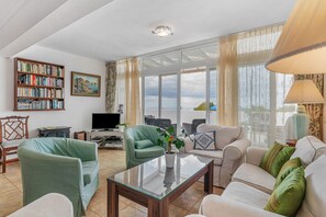 A living room with a sofa, chairs, a coffee table, a bookshelf, a TV, and a large window.