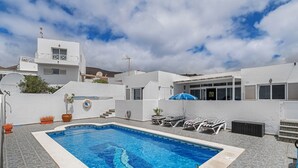 Una piscina con sillones de descanso y una zona cubierta, rodeada de edificios blancos y un cielo despejado.