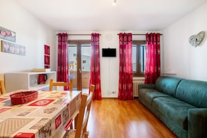Una habitación con una mesa de comedor, sillas, un sofá verde, cortinas rojas, un televisor y un reloj de pared.