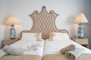 Uma cama de casal com cabeceira estofada, duas mesas de cabeceira com abajures e um cobreleite floral.