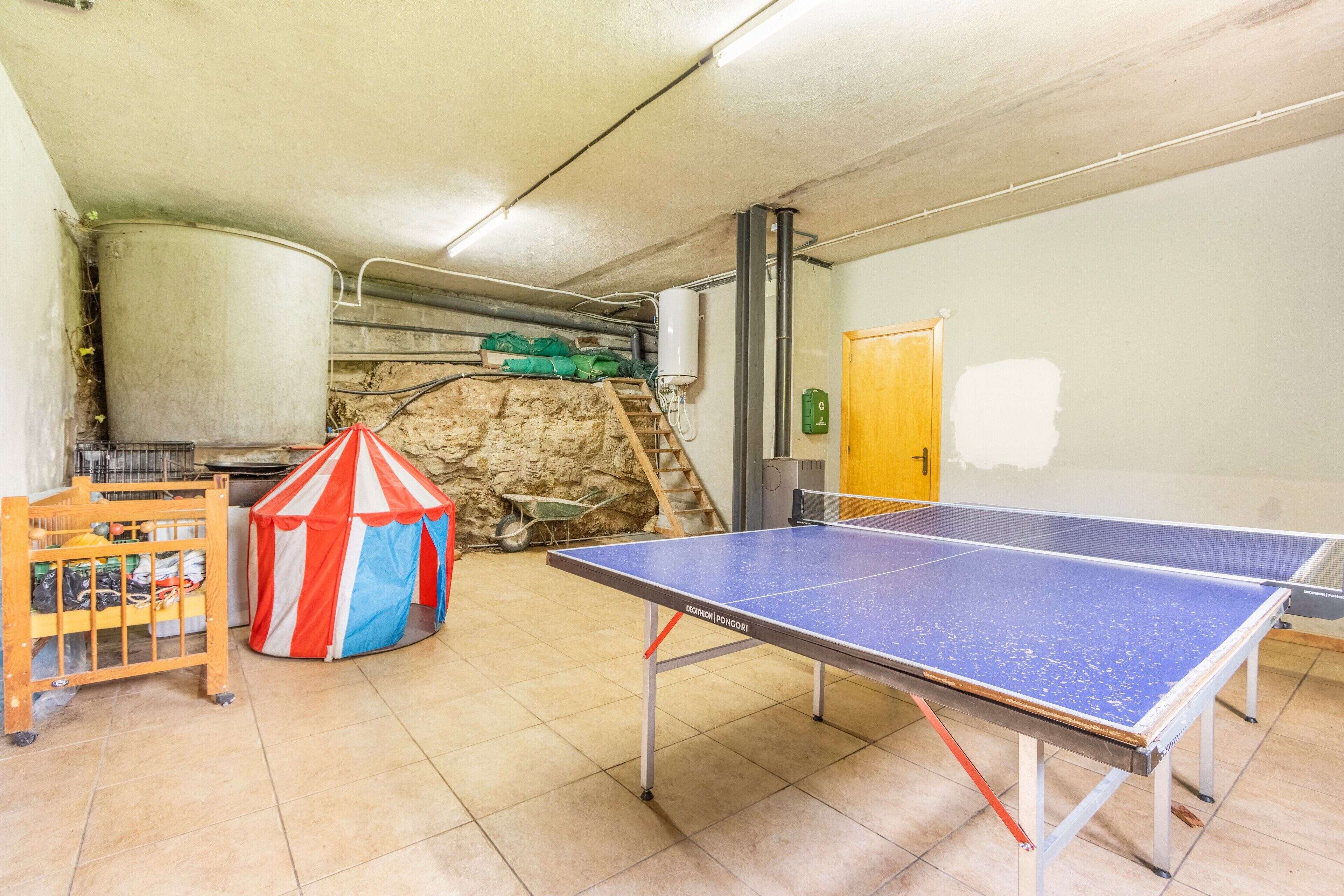 Una taula de ping-pong, una bressol de fusta i una tenda colorida en una habitació amb terra de rajola i paret de pedra.