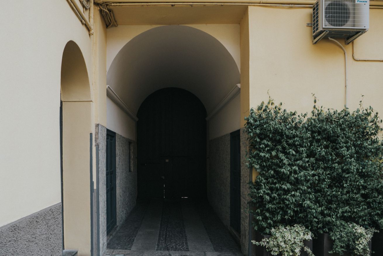 A corridor with an arched entrance, a wall-mounted air conditioning unit, and a hedge on the right.
