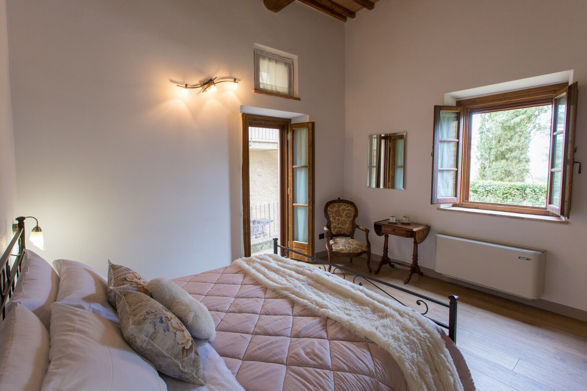Une chambre à coucher comprenant un lit, une chaise, une petite table, un radiateur et une fenêtre donnant sur un espace verdoyant.