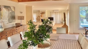A modern living room with a wooden ceiling, a dining area with white chairs, and a large potted plant.