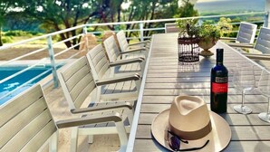 A wooden deck with outdoor seating, a bottle of wine, and a hat on a table, overlooking a pool and greenery.
