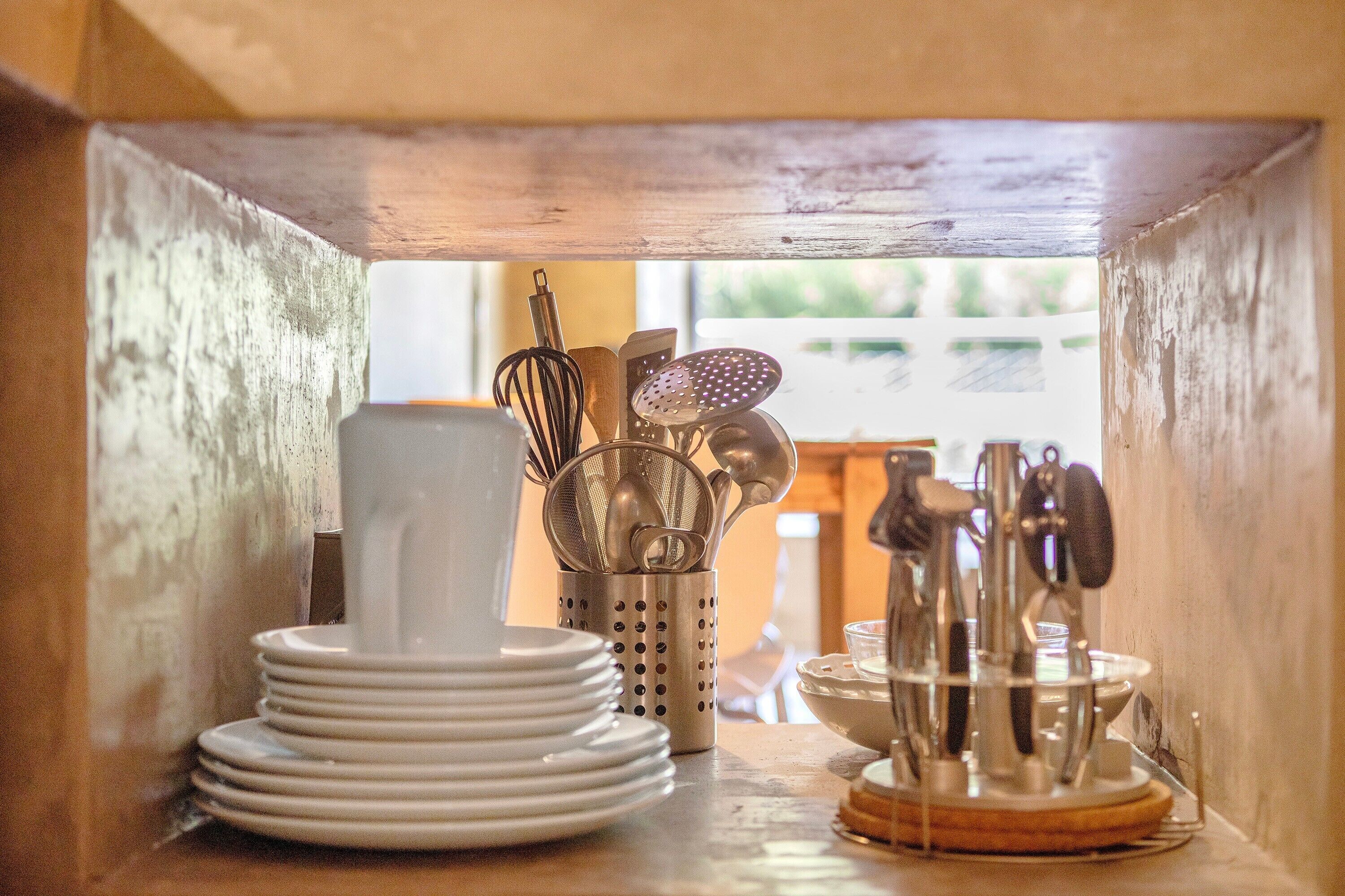 A kitchen area with a stack of white plates, a metal utensil holder, and a wooden cutting board.