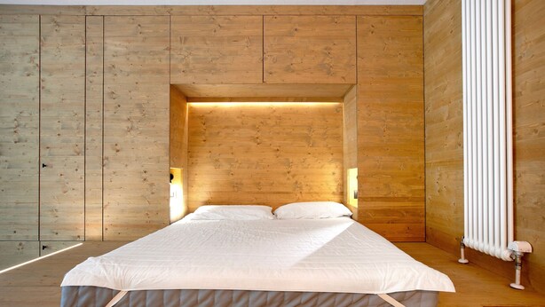 Une chambre avec un plancher en bois, un lit recouvert de draps blancs et un radiateur fixé au mur.