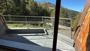 Un balcon doté d’une balustrade en métal, d’un sol en pierre et offrant une vue sur une chaîne de montagnes boisée.