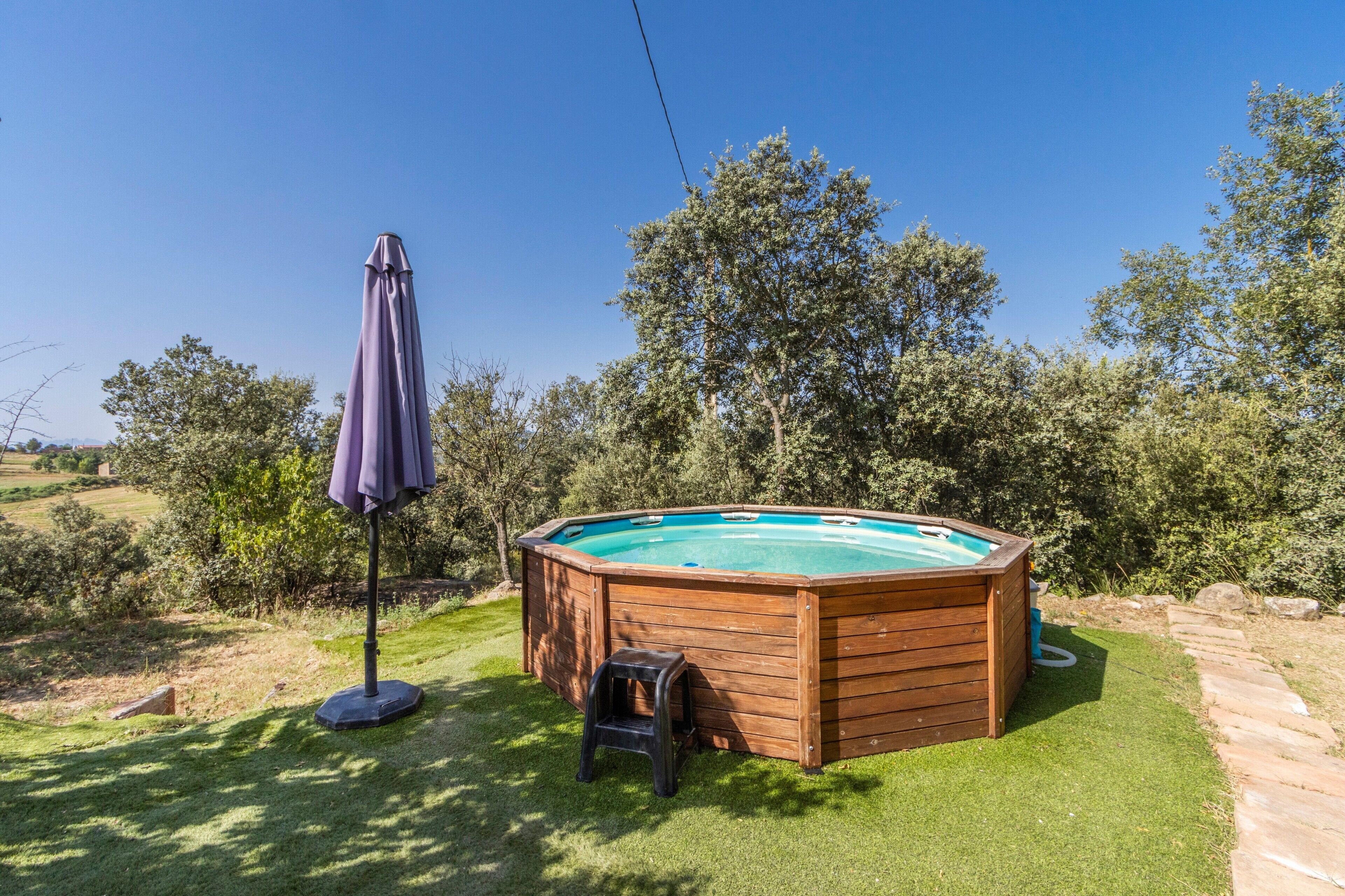 A wooden hot tub in a backyard with a lawn, a small stool, and a purple umbrella.