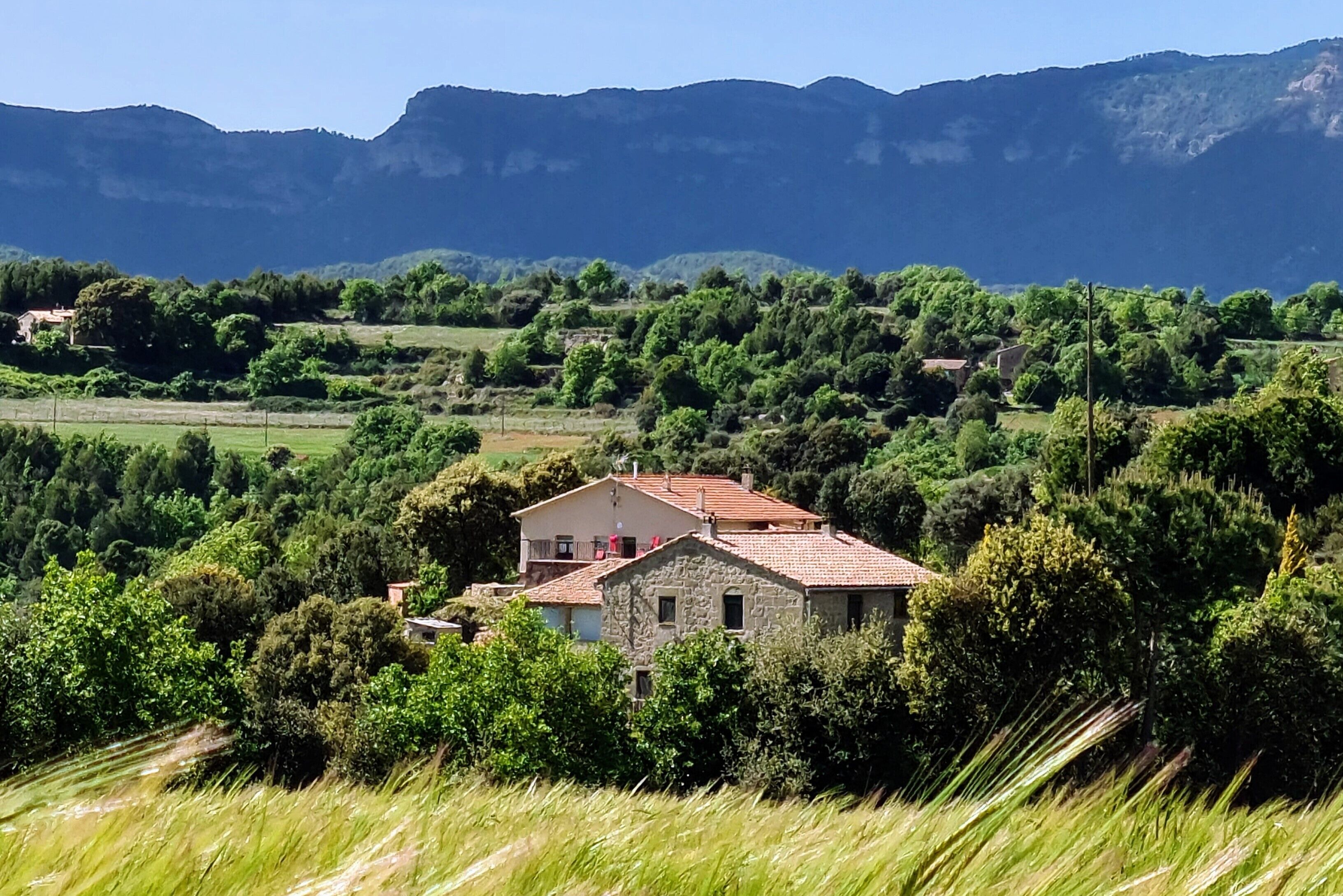 A stone house with a tiled roof surrounded by lush greenery and mountains in the background.