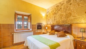 Quarto com parede de pedra, janela com persianas, radiador, cama com almofadas e mesa de cabeceira com abajur.